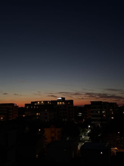 The image depicts a twilight cityscape with a gradient sky transitioning from dark blue at the top to orange and pink hues near the horizon. Silhouetted buildings, including a prominent multi-story structure with illuminated windows and the word "LUMA" in glowing letters on its facade, dominate the middle ground. Several other residential or office buildings are visible, some with lights on. The foreground shows darker, lower structures, possibly houses, also with some illuminated windows. Streetlights are visible, and a few vehicles are present on a street. The scene appears to be of a residential or mixed-use area in Năvodari, Romania, at dusk or early night. There are no visible people in the image.