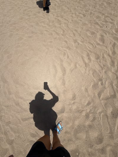 The image shows a view from above, looking down at a sandy surface, likely a beach in Badalona, Spain. In the foreground, a person's legs are visible, wearing black shorts and light blue sandals. The person is standing on the sand, with their shadow cast on the ground. The shadow indicates the person is holding a mobile phone, suggesting they are taking a picture or video. The sand is textured with ripples and small indentations, consistent with a beach environment. In the background, another person's legs and feet are visible, wearing dark shoes, with their shadow cast on the sand as well. The lighting suggests it is daytime, possibly with the sun casting distinct shadows. The overall scene captures a casual moment on a sandy outdoor location.