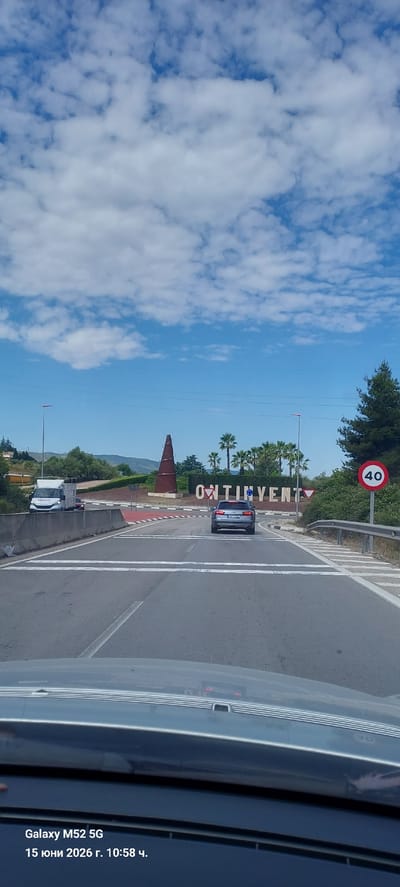 The image captures a view from inside a vehicle approaching a roundabout in Ontinyent, Spain. The sky is blue with scattered white clouds. In the background, large white letters spell out "ONTINYENT" on a raised circular island. To the left of the letters, a tall, reddish-brown, cone-shaped sculpture is visible. Several palm trees are planted around the roundabout, and to the far right, a circular speed limit sign indicates "40". A silver Audi car is in the lane ahead, approaching the roundabout. Further to the left, a white truck with a cargo box is visible. The road ahead has white lane markings and pedestrian crossing lines. The foreground shows the dashboard and windshield of the vehicle from which the photo was taken, with a timestamp in the lower left corner reading "Galaxy M52 5G 15 юни 2026 г. 10:58 ч.".