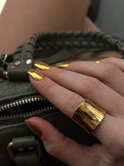 A close-up photograph shows a person's hand wearing a large, gold, statement ring on the middle finger. The fingernails are painted a matching metallic gold color. The hand is resting on what appears to be a green, leather handbag with a zipper and braided handle. The setting or location is not definitively conveyed in the provided image. There is no definitive evidence of Kerobokan Kelod, Indonesia, nor does the scene convey location information.