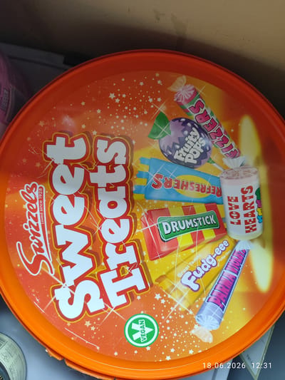 This is a close-up, overhead shot of the lid of a large orange tub of Swizzels "Sweet Treats" candy. The lid is decorated with colorful cartoon images of various sweets, including Drumsticks, Love Hearts, Fudg-eee, Fruity Pops, Refreshers, and Fizzers. The text "Swizzels Loved since 1928" is prominently displayed in white script on the left side of the tub. Large white block letters spelling "Sweet Treats" dominate the center of the lid. A green circular "VEGAN" logo is visible on the bottom left. The lighting is bright, suggesting it could be daytime, and the setting appears to be indoors, possibly a retail environment or a home pantry, given the background elements and the timestamp "18.06.2026 12:31" which suggests a date and time. No people are visible in the image. The overall mood is cheerful and playful, reflecting the nature of the candy products.