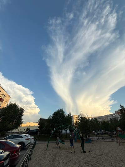 The image depicts a playground area in Surgut, Russia, under a dramatic sky filled with large, wispy clouds. In the foreground, a sand-covered playground features several children playing. One child, wearing a white cap and blue shorts, stands near a green swing set. Another child, wearing a dark shirt and white cap, is further back on the playground. A dark figure stands near the swing set. In the background, parked cars, including a white SUV and a red sedan, are visible along a fence line. Apartment buildings and trees form the backdrop of the scene. The sky is a vibrant blue, with the large white clouds dominating the upper portion of the frame, catching the warm light of what appears to be sunset.