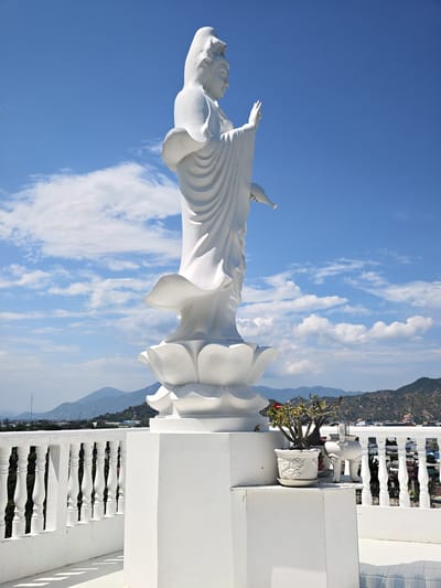 A full-shot, eye-level, outdoor photograph captures a large, white, ornate statue of a female deity, likely Guan Yin, standing atop a tiered lotus pedestal, which in turn rests on a tall, white, square base. The statue, illuminated by bright sunlight, faces to the right and is depicted with flowing robes and one hand raised in a blessing gesture, the other holding a vase. Behind the statue, a white railing with decorative balusters forms a border on a white platform. In the foreground, to the right of the statue's base, a small potted plant with red flowers and an incense burner are visible. The background reveals a panoramic view of the city of Phan Rang – Tháp Chàm, Vietnam, with distant, hazy mountains under a vibrant blue sky dotted with scattered white clouds. The overall scene suggests a religious or spiritual site offering a scenic overlook.