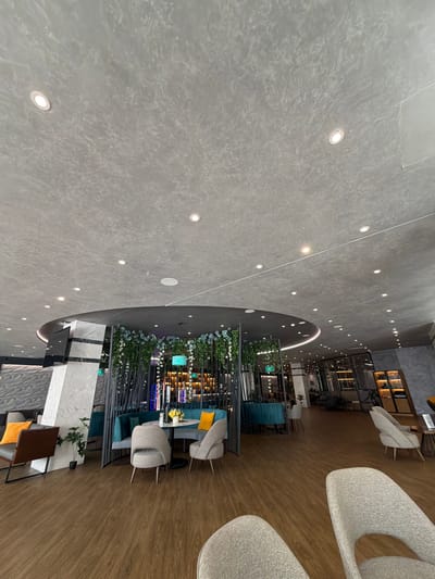 The image depicts a wide-angle view of a modern interior space, likely a cafe or lounge area, in Hisarya, Bulgaria. The scene is captured from a high vantage point, looking down and across the room. The flooring is a mix of textured grey material and polished wood.
The primary subjects are the seating arrangements. There are several comfortable-looking chairs with grey upholstered seats and backs, some with prominent curves. A few of these chairs are arranged around small tables. In the background, a bar or retail area is visible, featuring shelving units stocked with various items, illuminated by warm lighting. Decorative elements like artificial plants and flowers are strategically placed throughout the seating areas, adding a touch of greenery.
The lighting in the space appears to be a combination of recessed ceiling lights, which create a pattern across the grey ceiling, and warmer, more ambient lighting emanating from the background shelves and perhaps from within some of the decorative elements.
The time of day is not explicitly indicated, but the interior lighting suggests it could be evening or a dimly lit daytime environment. The overall atmosphere appears calm and inviting, with no people actively visible in the frame, suggesting a quiet moment or that the photo was taken before or after a busy period. The modern design, clean lines, and neutral color palette contribute to a sophisticated and contemporary feel. The location cues are limited to the provided context of Hisarya, Bulgaria. No visible text is present in the image.