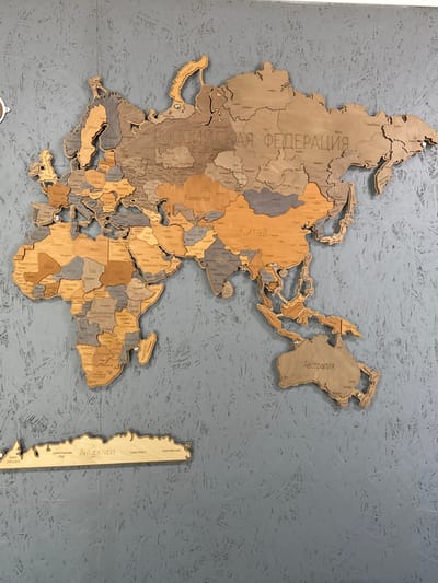 This media file, captured in Alamüdün, Kyrgyzstan, displays a large, decorative wooden world map mounted on a textured gray wall. The map is composed of numerous individual wooden pieces, varying in light and dark brown tones, representing continents and countries, which are slightly raised, creating a three-dimensional effect. Small metal fasteners secure some of the larger pieces to the wall.

The map's projection is stylized, with a significant focus on Europe, Asia, and Africa, appearing roughly centered. Australia is visible to the right, and Antarctica is presented as a separate, elongated piece on the far right. Many geographical entities are labeled directly on the wood in Cyrillic script.

Notable visible text includes: "РОССИЙСКАЯ ФЕДЕРАЦИЯ" (Russian Federation), "Казахстан" (Kazakhstan), "Китай" (China), "Монголия" (Mongolia), and "Австралия" (Australia). On the Antarctica piece, "Антарктида" (Antarctica) is prominently displayed, along with "Земля Королевы Мод" (Queen Maud Land), "Земля Элсуорта" (Ellsworth Land), "Земля Уилкса" (Wilkes Land), and "Земля Виктории" (Victoria Land). Other visible country names include "Украина" (Ukraine), "Беларусь" (Belarus), "Германия" (Germany), "Судан" (Sudan), "Египет" (Egypt), "Мали" (Mali), "Мадагаскар" (Madagascar), and "Демократическая республика Конго" (Democratic Republic of Congo). The gray wall has a rough, chipboard-like texture, suggesting it might be painted OSB or similar material. The scene is brightly and evenly lit, indicative of an indoor setting.