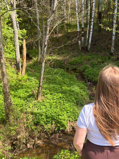 A person with long blonde hair is standing near a small, overgrown stream or ravine in a wooded area. The person is wearing a white shirt and brown pants. The area is covered in lush, green foliage and trees, including birch trees with white bark in the background. There is no specific evidence to conclusively indicate this image was taken in Ural'skoe, Russia. It is not possible to verify the location of the image with the information provided.