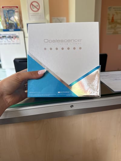 The image shows a close-up of a hand holding a box of Opalescence tooth whitening systems. The box is white with blue and silver accents, and features the Opalescence logo and brand name. The hand is well-manicured, with a pink nail polish. In the background, out of focus, are some items on a counter, possibly in a dental office or clinic, including a sign with a crossed-out cigarette symbol. The lighting suggests an indoor setting, possibly during daylight hours. The overall impression is a product presentation or advertisement. Visible text includes the product name "Opalescence™ tooth whitening systems", a phone number "800.552.5512", a website "ULTRADENT.COM", and manufacturing information.