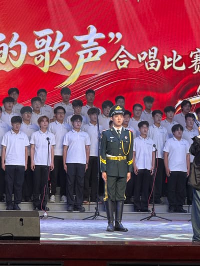 The image captures an indoor performance on a brightly lit stage in Guta, China. In the foreground, a prominent figure stands in a formal green Chinese military uniform adorned with gold trim and epaulets, a military cap, and tall black boots. Behind this individual, multiple rows of young men are arranged like a choir, primarily dressed in white short-sleeve polo shirts and dark trousers. Microphones on stands are visible amongst the performers.
The backdrop is a striking red, featuring large white and yellow Chinese characters. The visible text reads "唱红歌 颂祖国" (Chàng hóng gē sòng zǔ guó), which translates to "Sing red songs, praise the motherland." This strongly suggests a patriotic or commemorative event. The performers and the person in military attire appear focused and composed, contributing to a formal and solemn atmosphere. The artificial lighting indicates that the event is taking place indoors, likely in the evening.