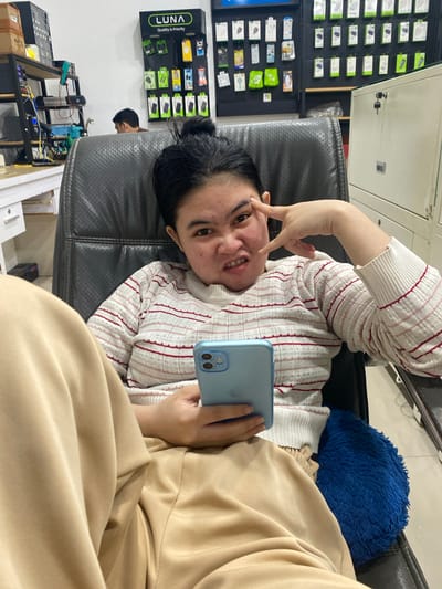 In the city of Ta', Indonesia, a young woman sits in a black leather office chair in what appears to be a store or workshop. She is wearing a white sweater with thin red and pink stripes and beige pants. She holds a light blue smartphone in her right hand and makes a playful gesture with her left hand, pointing towards her eye with her index finger and making a V-shape with her other fingers. Her facial expression is one of amusement. Behind her, a wall displays various phone accessories, including chargers and headphones, under a sign that reads "LUNA Quality is Priority." To the left, a shelf unit holds electronic equipment and boxes. A metal filing cabinet with a lock is visible on the right. Another person, partially obscured, is seated in the background. The environment suggests a retail or repair setting for electronics.
