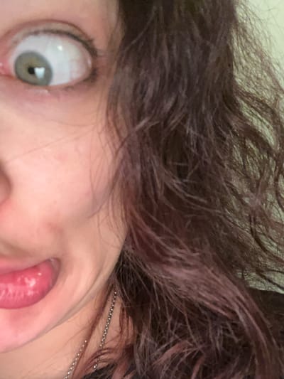 This is an extreme close-up portrait of a person's face, captured with a slightly tilted perspective. The subject has dark, wavy hair, appearing dishevelled, with hints of reddish-purple streaks visible on the right side.

The primary focus is on one wide-open eye, which features a light green or hazel iris, a dilated pupil with a prominent light reflection, and a significant portion of the white sclera exposed, contributing to a startled or exaggerated expression. Below the eye, part of the nose and cheek are visible. From the side of the mouth, a bright pink tongue is partially extended or pushed out, adding to the playful or quirky demeanor. The skin appears fair.

A delicate, silver beaded chain is visible, running through the hair on the upper right side of the frame. The lighting seems to be artificial, given the distinct reflection in the eye. The image conveys a sense of informal, spontaneous self-expression or a humorous moment. No text is visible in the image. The context provided indicates the location is Picassent, Spain.