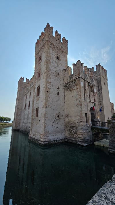 The image shows the Rocca Scaligera, a fortified castle located in Sirmione, Italy. The castle is situated on a small peninsula jutting into Lake Garda, and its stone walls are surrounded by water. The castle is a formidable structure with tall towers and crenellated battlements. Two flags, one Italian and one possibly another European nation, fly near the entrance. A bridge leads to the castle gate. The water surrounding the castle is dark and reflects the castle's structure. The sky above is clear blue with a few wispy clouds. The overall impression is one of historical grandeur and architectural resilience.