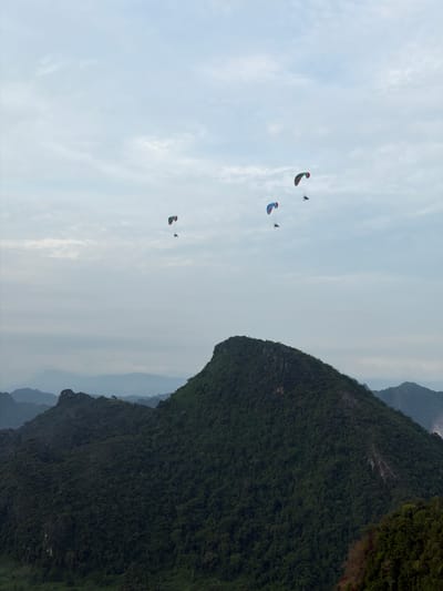 Four paragliders are visible in the sky above lush, tree-covered mountains in Vang Vieng, Laos. The paragliders, each with a colorful canopy and a pilot suspended below, are spread out across the frame. In the foreground, a large, dark green mountain dominates the landscape, its slopes densely covered in trees. In the background, several layers of smaller, blue-hued mountains recede into the distance, creating a sense of depth. The sky is a pale, overcast blue with wispy clouds.