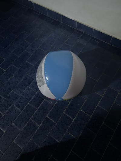 A slightly deflated, multi-colored beach ball rests on a dark blue tiled floor. The tiles appear to be arranged in a herringbone pattern. The wall is visible on the left edge of the scene. It's a sterile, indoor environment. Location unknown.