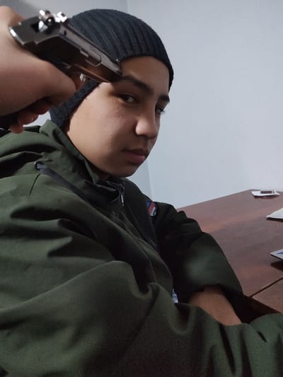 A person in a dark green jacket and a black beanie is looking at the camera while someone holds a pistol to their forehead. They are seated at a dark wooden table. A small object, possibly a card or piece of paper, rests on the table beside them. The wall behind is a light gray or white. The overall scene conveys a tense and potentially dangerous situation.