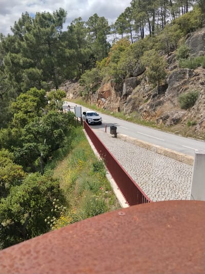 The image depicts a rural scene in Alijó, Portugal, on a sunny day with partial cloud cover. The viewpoint is elevated, possibly from a bridge or structure, overlooking a winding road that cuts through a landscape of trees, rocky terrain, and some vegetation. A white car is positioned on the road, heading towards the viewer. Adjacent to the road is a pedestrian walkway with cobblestone paving, marked by a reddish-brown railing. A trash receptacle is visible along the walkway. In the background, a steep hillside is covered with trees and rocks. In the distance, on the left-hand side of the car, two people are visible on the walkway. The foreground is dominated by a rusted metal surface.