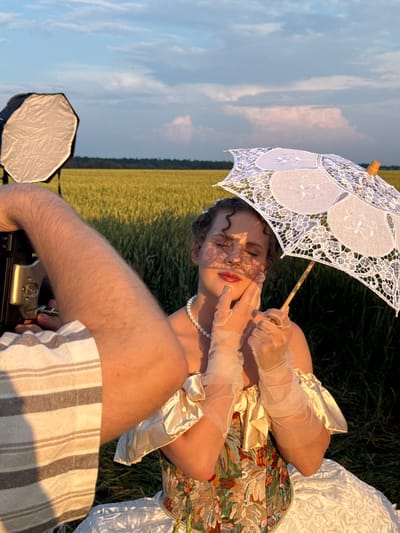 This is a medium shot photograph taken outdoors in a field of golden wheat under a cloudy sky. The scene is being filmed or photographed by someone whose arm and shoulder are visible in the foreground, holding a camera. A lighting diffuser is positioned to the left of the frame.

The primary subject is a person, appearing to be female, with dark curly hair, dressed in an ornate, vintage-style gown with a corseted bodice featuring a floral pattern and a voluminous white skirt. She is wearing sheer, elbow-length gloves and a pearl necklace. Her eyes are closed, and she is holding a delicate white lace parasol above and to the right of her head. Her hands are positioned near her face, with one hand touching her cheek. The sunlight casts a dappled pattern on her face through the lace of the parasol. The background consists of the wheat field and a treeline in the distance. There is no visible indication of the city of Foki, Russia, in this image.