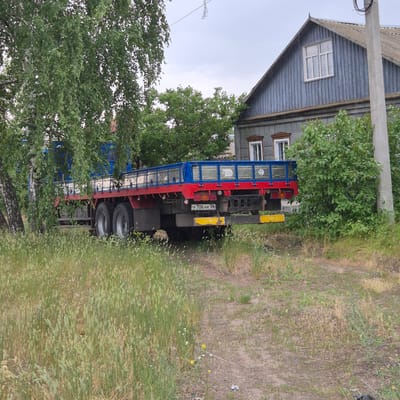 In the city of Liski, Russia, a flatbed truck with a red chassis and a blue railing is parked on a dirt road next to a grey wooden house. The truck is partially obscured by trees and tall grass. The house has a grey metal roof and blue wooden siding, with two windows on the upper floor and two smaller windows on the lower floor. A concrete utility pole stands to the right of the house. The truck's license plate is visible and reads "У 706 НК 36". The scene is set in a rural or semi-rural environment under an overcast sky.