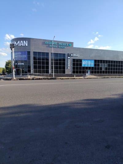 A wide shot captures a modern, two-story commercial building with a grey tiled facade under a bright blue sky with scattered white clouds. The building is situated at an intersection with a traffic light visible on the left. The building features large, dark-tinted windows that reflect the sky. Prominent signage in both Russian and what appears to be Kazakh adorns the facade. On the left, large white letters spell out "OPMAN". To the right of "OPMAN", a blue sign reads "ЦЕНТР МАТРАСОВ" (Center of Mattresses), with smaller text below it. Further to the right, teal lettering on the building states "АКВОРЕ MEBEL" (Aquore Furniture), with smaller text below. Another sign to the far right reads "ЖИНАЗ" in teal letters with a logo, and vertical text spelling out "МЕБЕЛЬ" (Furniture) in black on the left side of this section. A blue illuminated sign displaying text related to mattresses and furniture is visible in a window to the right. A sidewalk runs in front of the building, with a ramp and railings leading up to an entrance. The foreground shows a paved road with a shadow cast across it, suggesting it is daytime. The environment appears to be an urban or suburban setting.