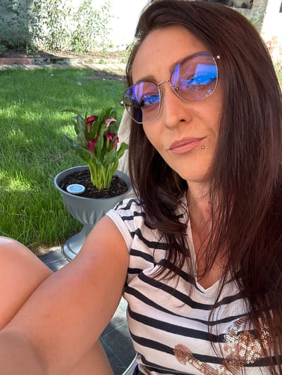 A woman is taking a selfie outdoors in Villa Pasquali, Italy. She has long brown hair and is wearing large, reflective sunglasses and a black and white striped shirt with sequin embellishments. She is sitting on a mat in front of a grassy area and a potted plant with dark red calla lilies. The sun is shining, casting shadows and reflecting off her sunglasses.