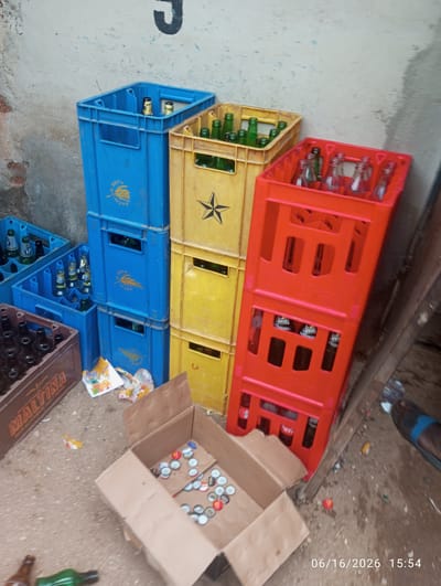The image depicts an outdoor scene, likely in Dokan Tofa, Nigeria, characterized by the presence of multiple plastic crates filled with bottles. In the foreground, a brown cardboard box is open, containing numerous bottle caps of various colors. To the left of the box, a brown wooden crate labeled "MALTINA" is visible, also containing bottles. Further into the scene, several plastic crates are stacked and arranged: a blue crate is positioned to the left, a yellow crate in the middle, and a red crate to the right, all holding bottles. Scattered on the ground are more bottles, including a green one in the lower left and a brown one next to it. The ground appears to be a mixture of concrete and dirt, with debris present. In the upper left corner, a portion of a wall with some painted markings is visible. The overall impression is one of an accumulation of beverage containers and caps in an outdoor setting.