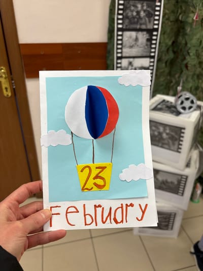 A right hand, likely Caucasian and adult, holds a handmade greeting card in the lower-left foreground. The card features a light blue background depicting a sky, with a three-dimensional hot air balloon constructed from paper segments colored white, blue, and red, mirroring the Russian national flag. A yellow paper basket hangs below the balloon by drawn brown lines, displaying the number "23" in orange-red. Three white paper clouds are affixed to the blue background. Below the central image, the word "Fевruary" is written in red marker, utilizing a Cyrillic 'в' character.
The background shows an indoor environment. To the left, a segment of a light brown wooden door with a gold-colored doorknob and lock mechanism is visible. To the right, decorative elements are arranged. These include a vertical strip resembling a film reel, containing multiple black-and-white photographic images, some appearing to depict military or historical scenes. Below this strip, several white cubic boxes are stacked, each decorated with similar black-and-white film-strip images. A silver-colored object resembling a film reel spool rests atop one of these boxes. A green, textured fabric or netting is partially discernible behind the decorations. The floor is covered with light-colored tiles and dark grout. The scene is located in Novosergievka, Russia.