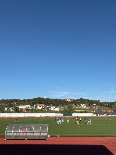 The image depicts a soccer match in progress on a green field, alongside a red running track, under a clear blue sky. A hillside with houses, trees, and structures, including a tower, flanks the field. On the sideline, a team dugout is visible. Several soccer players in blue and white uniforms are grouped on the field, suggesting a potential play or set piece. The location is likely Valpaços, Portugal, based on the provided contextual information.
