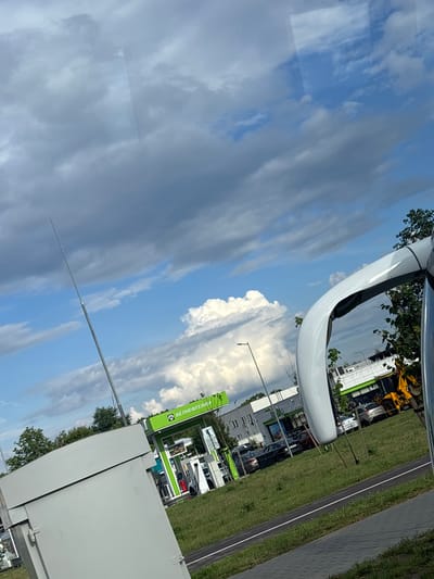 The media file is a photograph taken outdoors, featuring a bright sky with scattered clouds. The image is oriented vertically. In the foreground, a portion of a large, white, curved object is visible, possibly part of a vehicle or a structure.
The background reveals a street or road with a grassy verge. Beyond that, there are buildings, some with green awnings or signage. A yellow construction vehicle is partially visible among the buildings. Trees are scattered throughout the scene.
A prominent green sign with white lettering is visible. The text on the sign appears to be in Cyrillic script and reads "БЕНЕКОТЕХНИК" and below it, "ВАСИЛЕВ".
The lighting suggests it is daytime, likely a sunny day given the brightness of the sky and clouds. The weather appears to be clear with some cloud cover. The location context provided indicates the setting is within the Senitsa village Council in Belarus. There are no people clearly visible in the image. The overall scene depicts an outdoor commercial or industrial area.