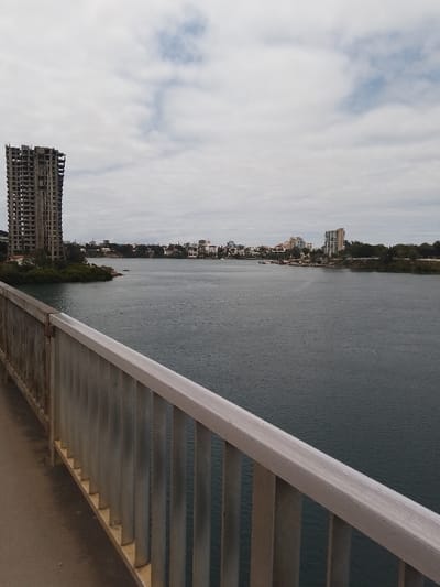 The image depicts a panoramic view across a body of water in Junda ward, Kenya, observed from a bridge or elevated structure under an overcast sky. In the foreground, a bridge railing composed of a horizontal beige-colored top rail and vertical gray metal pickets extends diagonally from the lower right towards the mid-left. A dark gray pathway surface is visible adjacent to the railing on the left. The midground features a wide expanse of dark blue-green water, exhibiting minor surface ripples. Along the left bank, a prominent unfinished concrete high-rise building, approximately fifteen stories tall with exposed structural elements and numerous open window frames, stands amidst dense green foliage. Further along this shoreline, a cluster of various white and light-colored multi-story urban buildings is visible. The right shoreline also presents urban development, including several completed multi-story buildings and interspersed green trees. Small boats or watercraft are discernible on the water near both shorelines. The sky is covered by diffuse gray and white clouds, with occasional lighter blue patches visible. No people or specific actions are visible within the scene.