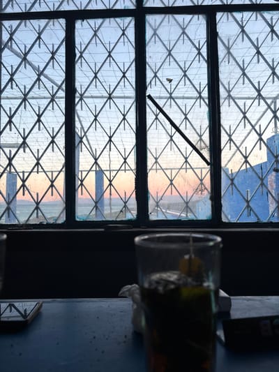 A window with a diamond-shaped metal grate overlaying the glass panes dominates the image. The view through the window is a mix of sky and a horizon line with warm colors. To the right, a glass containing a dark substance is visible, along with other blurry objects suggesting a cluttered shelf. The entire scene is rotated, presenting a sideways view of the interior.