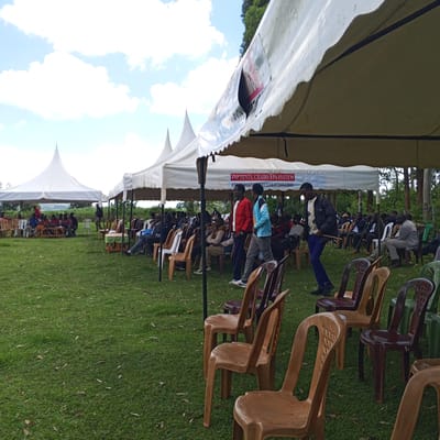 The image shows an outdoor gathering in Keyian ward, Kenya. Several white tents are erected on a green lawn, providing cover for seated individuals. Numerous plastic chairs in varying colors (brown, maroon, green) are arranged in rows facing a group of people seated in the distance, possibly at the front.
People are seen sitting and standing around the area. Some are casually dressed and walking between the rows of chairs.
The backdrop includes trees and foliage, suggesting a rural or semi-rural setting. The sky is partly cloudy.
