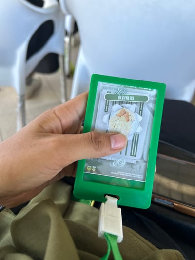 A person's hand is holding a green ID card holder containing an ID card with a picture of a person wearing a white shirt and what appears to be a white head covering. Text on the ID card includes "SRI RAHAYU" and "ANITA". The ID card is within a green plastic holder. The background is blurred, showing white chairs, suggesting an indoor setting, possibly a waiting area or classroom. The person's clothing is visible at the bottom of the frame, appearing to be olive green fabric. The location is not specified in the image.