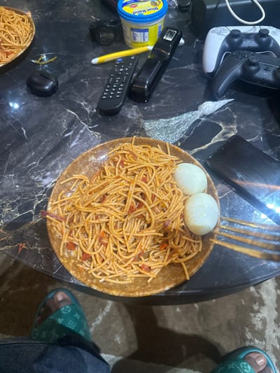 The scene is indoors, in Orie Uratta, Nigeria, viewed from a first-person perspective looking down at a dark, polished, possibly marble-patterned table surface. The floor beneath the table is covered with a textured, light brown carpet.
On the table, a brown, speckled plate holds a serving of spaghetti mixed with a reddish sauce containing diced red and purplish ingredients, likely tomatoes and onions. Two peeled hard-boiled eggs rest on the right side of this plate. A second, partially visible plate of similar spaghetti is positioned in the upper left corner of the frame.
Other objects on the table include a blue and yellow tub of "Blue Band" margarine, a black remote control, a yellow pen, and a black stapler. To the right, a white PlayStation 5 DualSense controller is stacked upon a black PlayStation 5 DualSense controller. A tangled white cable is visible near the controllers. A black mobile phone lies face down to the right of the main spaghetti plate. Further left, a black wireless earbud case is present. A small, crumpled piece of transparent plastic film is on the table between the remote and the mobile phone. A metallic fork is partially visible to the right of the eggs.
The lower portion of a person is visible, wearing blue denim jeans and light green/teal open-toed slides with a repeating, light-colored pattern, indicative of a luxury brand. The person's left foot is clearly visible, with the toes exposed, and a portion of their right foot is also visible.