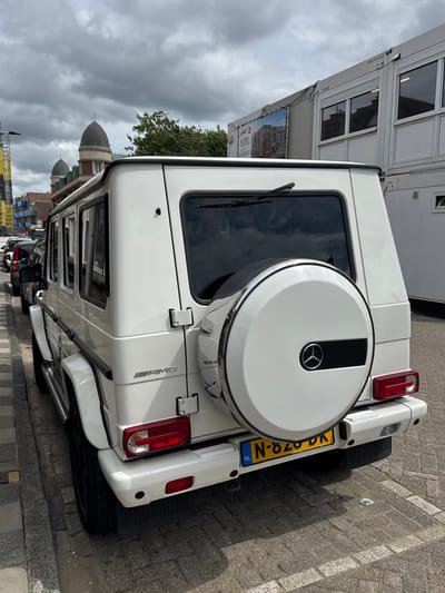 A white Mercedes-Benz G-Wagen, identifiable as an AMG model by its prominent badge on the rear passenger side panel, is the central subject of the image. The vehicle is seen from an elevated perspective, showcasing its rear and passenger side as it sits parked on a paved surface. The spare tire, enclosed in a white cover bearing the Mercedes-Benz logo, is mounted on the tailgate. A Dutch license plate, "N-820-DK," with the "NL" country identifier, is clearly visible on the rear bumper. Other visible details of the car include the rear lights, a rear windshield wiper, and side running boards.
The setting consists of grey and tan cobblestone paving where the car is parked. In the background, several white modular container buildings are stacked, some featuring windows that reflect the cloudy sky. Text like "containerbouw" and "9805" is discernible on one container, while another displays a large printed image of a building. Beyond these structures, green tree foliage and distinct architectural elements with domed roofs are visible, set against a heavily overcast sky with significant cloud cover. The scene depicts a quiet, daytime moment with no people present, and the vehicle appears to be stationary. The location context provided is Noordwijk, Netherlands.