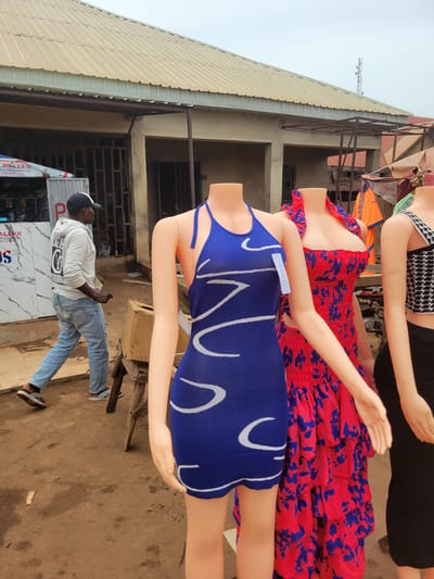 This image captures an outdoor market scene in Bokkos, Nigeria. In the foreground, three headless mannequins are displayed. The mannequin on the left wears a blue halter dress with white abstract patterns. The middle mannequin is adorned in a red and blue floral print dress with ruffled layers. The mannequin on the right is dressed in a black and white patterned top and a black skirt. Behind the mannequins, a man wearing a white hoodie, blue jeans, and a black and white cap walks away from the camera, his gaze directed to the right. He is wearing grey and white sandals. The market appears to be set up under a corrugated metal roof supported by metal poles. To the left of the mannequins, a stall is visible with various items displayed, including a cooler and signage. The ground is a mixture of dirt and concrete. The overall environment suggests a bustling, open-air marketplace typical of a Nigerian town.