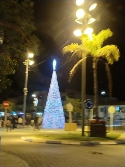 At night, a brightly lit artificial Christmas tree stands tall in the center of a roundabout. Streetlights illuminate the scene, with a palm tree visible to the right of the Christmas tree. Blurred figures of people walking by can be seen in the background. Traffic signs surround the roundabout, and buildings with illuminated windows are visible further back. The photograph is slightly blurry.