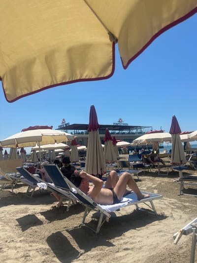 The image depicts a beach scene in Bashkia Durrës, Albania. A man is lying on a beach chair on the sandy beach, partially under an umbrella. Multiple rows of umbrellas and beach chairs extend into the background, with a building labeled "PORTO VIVO" visible further back, possibly a beachside establishment. The sky is clear and blue, indicating a sunny day.