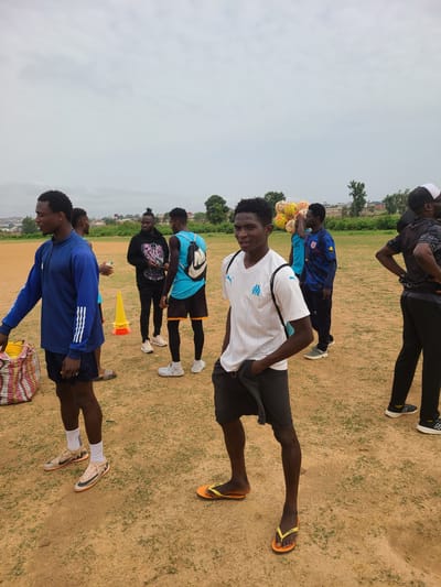 The scene is an outdoor field in Barikin Jirga Summa, Nigeria. A group of at least eight young men are present. They appear to be associated with soccer or sports, as evidenced by several soccer balls and training cones present. The environment is a grassy, dirt field, with trees and buildings visible in the background. The sky is overcast.

Several of the individuals are wearing athletic attire, including soccer jerseys and shorts. One man in the foreground is wearing a white jersey with the Marseille logo. Some are stretching or warming up. One individual is holding a bunch of soccer balls. The attire of the people suggests a warm climate.