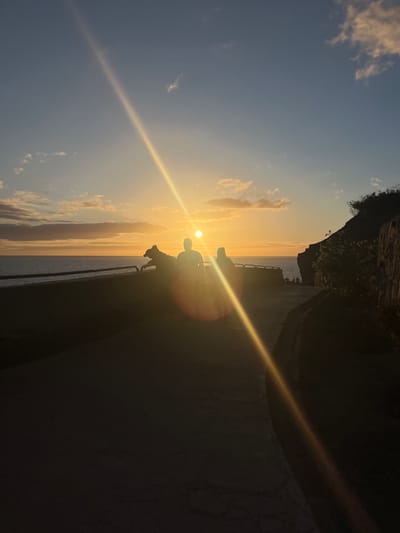 The image shows a sunset over the ocean, likely taken in Puerto Rico, Spain. A silhouette of a person or people is visible on a ledge or wall in the foreground, potentially sitting or standing. The sun's rays are prominent, causing a lens flare. The sky is a gradient of blue to orange, with some scattered clouds. A dark cliff or vegetation is visible on the right side of the frame. The perspective of the image is rotated.