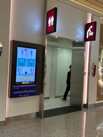 The image captures a modern public restroom entrance on the 5th floor, Concourse C, of Taiwan Taoyuan International Airport in Taoyuan City, Taiwan. A prominent vertical digital display board on the left wall indicates the real-time availability of restroom stalls, showing "Female" (女) with 5 available stalls and "Male" (男) with 3 available stalls, alongside icons and "Available" text. Below this, the screen displays information for a "Breastfeeding Room" and a directional map highlighting "5F" and "C6". Above the restroom entrance, two rectangular, illuminated red signs with white pictograms are affixed to the wall; one depicts male and female figures, and the other shows a baby changing symbol. The entrance itself features reflective, mirror-like surfaces. Inside the restroom, the floor has a small black and white hexagonal tile pattern, and the walls are light-colored. A single individual, dressed in a dark jacket, dark pants, and a dark baseball cap, is partially visible from the back and right side, standing within the reflective restroom entrance. The floor outside the restroom is a light gray speckled terrazzo. Vertical LED strips illuminate the wall sections surrounding the entrance.