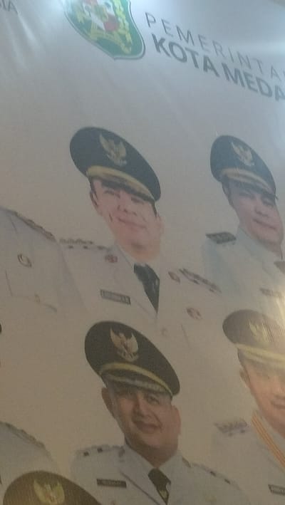 This media file features a banner with portraits of men in uniform, likely officials from the City of Medan, Indonesia. The banner displays the text "PEMERINTAH KOTA MEDAN" which translates to "Government of Medan City." The individuals are wearing dark caps with gold trim and a crest, and light-colored uniforms with dark ties. Each portrait appears to be a formal headshot. The overall impression is of an official display related to local governance in Medan. The lighting suggests an indoor setting or an overcast day, as there are no strong shadows. The style of the portraits and the banner suggests a public announcement or a display of leadership.