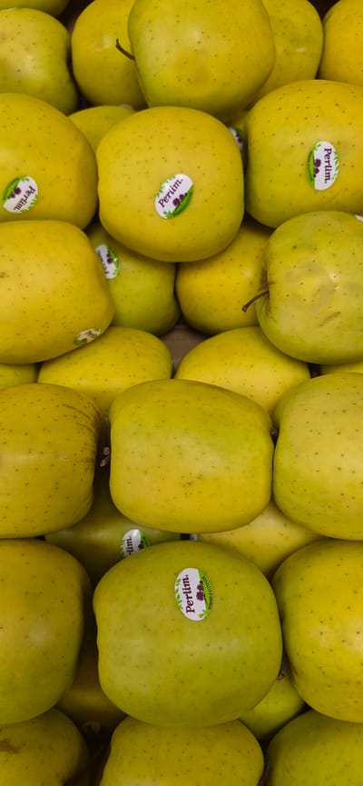 The image is a close-up, overhead shot of a pile of bright yellow-green apples, likely in a market or grocery store. The apples are stacked and appear to be whole and fresh. Many of the apples have small, round stickers on them that say "Perlim" and "POMME D'ALTITUDE". The lighting is bright and even, suggesting an indoor setting. The background is filled with more apples. The scene is sterile and does not provide any information about the city of Navalcarnero, Spain.