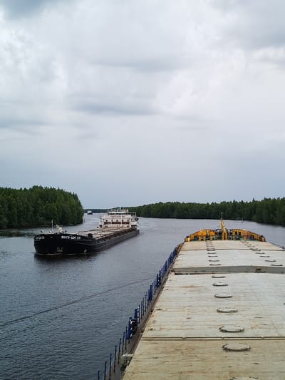 The image captures a wide shot of a river with two barges and a treed shoreline. In the foreground, the deck of a large barge, made of dull metal with circular hatches, stretches across the frame. The barge is painted blue on the sides. In the mid-ground, another barge, named "ВОЛГО-ДОН 139," is moving towards the viewer. This barge has a dark hull with "ВОЛГО-ДОН 139" written in white lettering and a white superstructure. A second, smaller barge is visible in the distance, further up the river. The riverbanks are densely covered with green trees, indicative of a natural, possibly rural, setting. The sky is overcast with grey clouds, suggesting a cloudy or potentially rainy day. The overall scene depicts maritime transport on a river in Novokemskii, Russia, under a subdued sky.