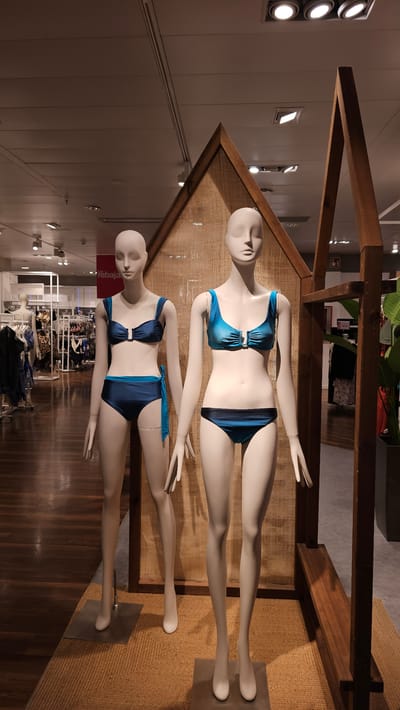 Two female mannequins are displayed in a retail store, likely a department store or clothing boutique. The mannequins are positioned in front of a backdrop with a textured, woven material and a wooden frame resembling a rustic shelter. Both mannequins are wearing bikinis. The mannequin on the left wears a dark blue bikini with a halter top and high-waisted bottoms that tie at the side. The mannequin on the right wears a metallic teal bikini with a scoop-neck top and matching bottoms. The floor is wooden, with a woven rug beneath the mannequins. In the background, racks of clothing and store fixtures are visible. The overall lighting suggests an indoor retail environment. This scene could be in Zaragoza, Spain, given the Spanish text "rebaja" (sale) visible on a sign in the background.