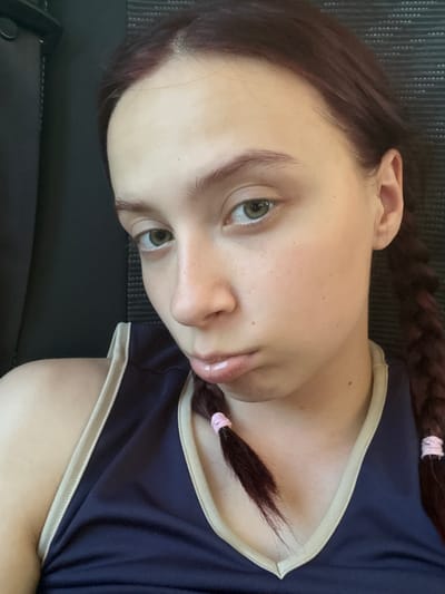 A medium shot depicts a young woman with fair skin, light green eyes, and reddish-brown hair styled in two braids secured with pink hair ties. She is wearing a navy blue top with a beige trim around the collar. The background is a dark gray, seemingly textured surface. The woman is looking towards the camera. The location in the city of Керамик, Russia is not discernible from the image.