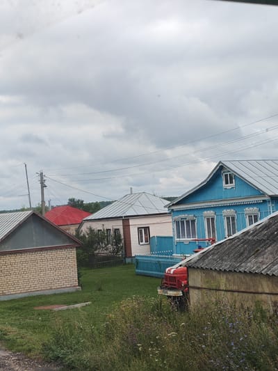 The image shows a cluster of rural houses in Yambirno, Russia. The foreground features a patch of overgrown grass with wildflowers. To the left is a light yellow brick house with a grey sloped roof. Adjacent to it, a house with a bright red roof is visible. Further back, a light-colored house with a white trim and a dark-pitched roof stands next to a vibrant blue house adorned with white decorative trim around its windows and roofline. In front of the blue house, a red fire truck is parked, partially obscured by a grey, weathered structure in the immediate foreground. Power lines traverse the cloudy sky above the scene.