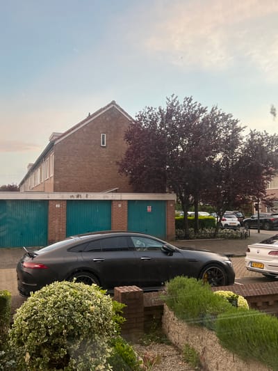 The image shows a street scene in Beverwijk, Netherlands. A matte black car is parked on the right side of the frame, partially obscured by a bush. To the left of the car are several garage doors, painted teal with brick accents. A brick building with a single, narrow window is visible above the garages. A tree with dark red foliage stands in front of the building. In the background, other parked cars are visible along the street. The sky is a pale blue with some cloud cover. The scene appears to be taken from inside a building looking out a window.