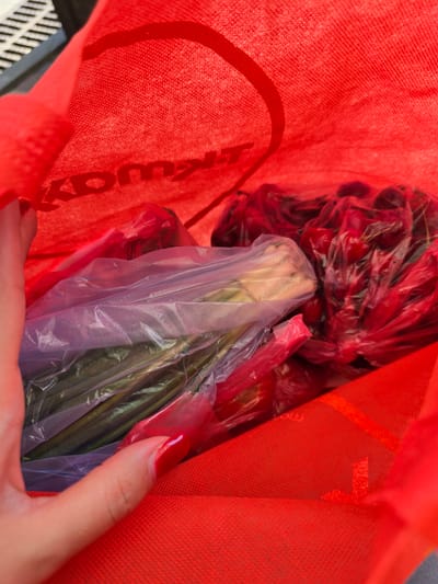 A hand with red fingernail polish is reaching into a red tote bag filled with produce. The tote bag has a logo visible on the inside, partially obscured by the contents. Inside the bag, there are clear plastic bags containing vegetables and red fruit. One plastic bag holds what appears to be green onions or a similar vegetable, while another bag contains red cherries or another red fruit. The scene is indoors, as indicated by the lighting and the presence of a shopping bag. The location is Petropavlivska Borshchahivka, Ukraine.