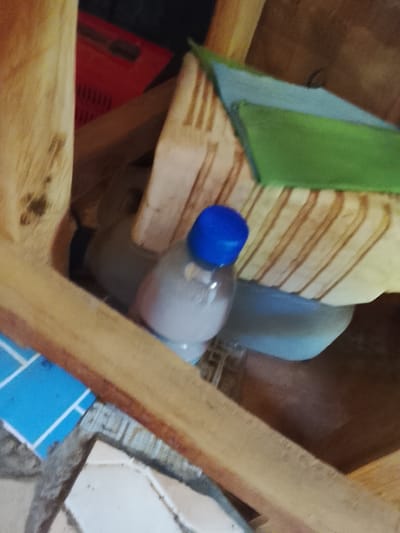 The image depicts a close-up, angled view of items within what appears to be an indoor storage area, possibly a corner of a room or a closet. The dominant materials are wood, plastic, and fabric. In the foreground, a wooden chair or shelf is visible, its unfinished planks forming structural elements. Resting on or near this wooden structure are several objects. A partially visible clear plastic bottle with a blue cap is in the center-right of the frame, containing a cloudy white liquid. Behind the bottle, a pale blue plastic container or bag is discernible, and further back, a bright red plastic crate with a ribbed texture is partially visible against a dark background. A piece of green fabric, possibly a cloth or mat, is draped over some of the wooden elements, and a light blue surface with a white grid pattern is in the lower left corner. The overall lighting suggests an indoor environment with ambient light. The image does not provide any specific visual cues to indicate its location within Sunkani, Nigeria.
