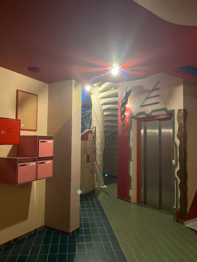 The image depicts an indoor scene that appears to be a lobby or common area, possibly in an apartment building or office complex. The orientation of the image is rotated, showing the ceiling and walls from a tilted perspective.

Prominently featured are several pink mailboxes stacked in a corner. To the left of the mailboxes, there's a corkboard with a red frame, and below that, a red fire alarm box. The walls are painted in varying colors, including a deep red on the left and a lighter, textured beige on the right. A section of the wall is covered in green and blue square tiles, characteristic of bathrooms or kitchens.

Further into the scene, a spiral staircase is visible, with some decorative elements, including what looks like a metallic sculpture resembling a dragonfly or butterfly illuminated by a bright light source. Above the staircase, there's a section of wall with abstract green lines and what appears to be an elevator door with a metallic finish, bordered by red and green trim. The overall aesthetic suggests a somewhat retro or artistic design.

The lighting in the scene is varied, with a strong bright light emanating from near the dragonfly sculpture, casting shadows and highlights. The time of day is not discernible, but the indoor setting and artificial lighting indicate it's likely not daytime. There are no people visible in the image. No text is legible on the mailboxes or any other visible surfaces. The location context is Geneva, Switzerland, but there are no specific visual cues within the image to confirm this.