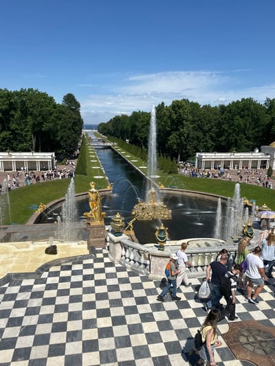 The image shows a panoramic view of the Grand Cascade at Peterhof Palace in Peterhof, Russia, on a clear, sunny day. The foreground features a black and white checkered tiled pathway, with several people walking on it. Beyond the pathway, a large fountain with golden sculptures and water jets is visible, with a man in a white t-shirt and shorts walking towards it. Further back, a long, rectangular canal stretches into the distance, flanked by manicured lawns, rows of trees, and smaller fountains. People are scattered throughout the scene, walking on pathways, and observing the fountains and architecture. In the background, a large body of water, likely the Gulf of Finland, is visible, as are the roofs of buildings, and more trees. The sky is a clear blue with a few wispy clouds.